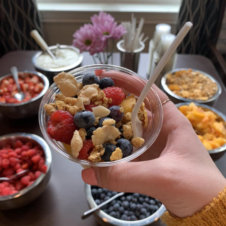Hand holding a yogurt parfait cup filled with fresh berries, granola, and crunchy toppings, with various fruit and granola bowls arranged on a table in the background.